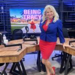 The Being Tracy Show - Smiling woman in a blue jacket and red dress stands in a podcast studio with a screen displaying "Being Tracy" behind her.