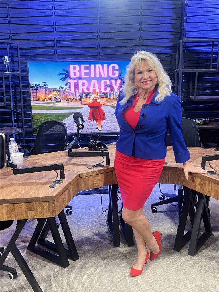 The Being Tracy Show - Smiling woman in a blue jacket and red dress stands in a podcast studio with a screen displaying "Being Tracy" behind her.