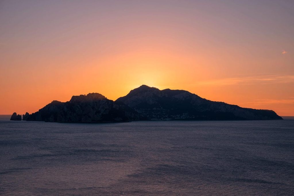 The Being Tracy Show - Island silhouette at sunset, with a vibrant orange sky and calm ocean in the foreground.