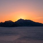 The Being Tracy Show - Island silhouette at sunset, with a vibrant orange sky and calm ocean in the foreground.