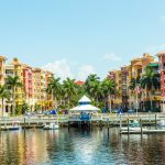 Colorful waterfront buildings with palm trees, boats, and a gazebo reflected on calm water under a clear blue sky.