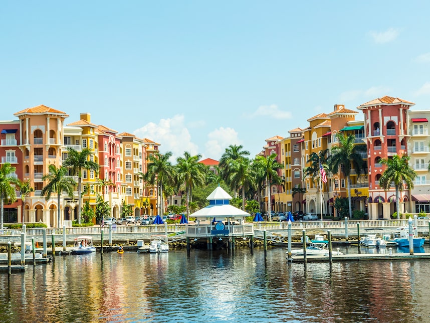 Colorful waterfront buildings with palm trees, boats, and a gazebo reflected on calm water under a clear blue sky.